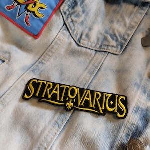 May include: A black embroidered patch with the band name "Stratovarius" in yellow lettering. The patch features a small fleur-de-lis design below the band name.