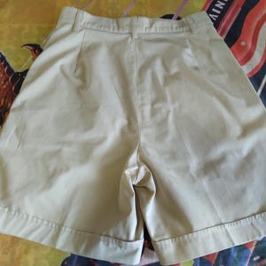 Puede incluir: Pantalones cortos de color caqui con la parte delantera plisada y un ajuste a medida. Los pantalones cortos tienen cierre de bot&oacute;n y cremallera, trabillas para el cintur&oacute;n y dobladillo con vuelta. La tela parece ser de algod&oacute;n ligero o mezcla, adecuada para climas c&aacute;lidos.