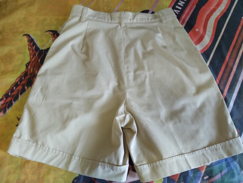 Puede incluir: Pantalones cortos de color caqui con la parte delantera plisada y un ajuste a medida. Los pantalones cortos tienen cierre de bot&oacute;n y cremallera, trabillas para el cintur&oacute;n y dobladillo con vuelta. La tela parece ser de algod&oacute;n ligero o mezcla, adecuada para climas c&aacute;lidos.