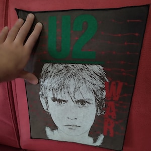 May include: A black patch featuring the band U2's logo in green and the word "WAR" in red. The patch has a black border and a grayscale image of a child's face.