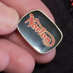 May include: A black and gold pin with the word "Norton" in red letters.
