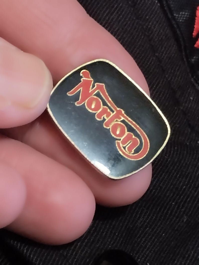 May include: A black and gold pin with the word "Norton" in red letters.