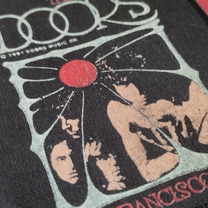 May include: A rectangular black fabric patch featuring a white graphic design with the text THE DOORS and 1991 DOORS MUSIC CO. A central red circle sits above stylized silhouettes of figures. The bottom edge displays the text FRANCISCO 19.