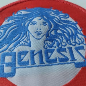 Könnte beinhalten: Ein kreisförmiger bestickter Aufnäher mit rotem Rand und weißer Mitte. Das Design zeigt eine blaue Stickerei einer Person mit langem, fließendem Haar und das Wort "genesis" in blauen Buchstaben unterhalb des Gesichts.
