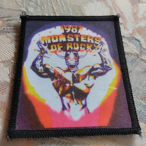 Puede incluir: Un parche de tela rectangular con un borde negro cosido que presenta un gráfico de una figura con cuernos y brazos levantados. El texto 1987 MONSTERS OF ROCK está impreso en amarillo y blanco sobre la figura, con un fondo morado y naranja.