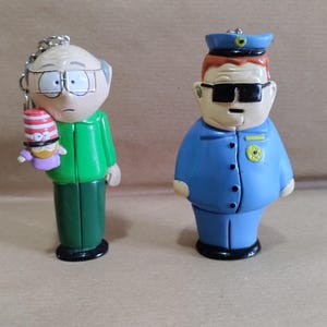 Puede incluir: Dos figuras de personajes de South Park. La primera es una figura verde de Mr. Garrison sosteniendo una pequeña figura roja y blanca de un personaje con un sombrero. La segunda es una figura azul de Officer Barbrady con un uniforme azul y gafas de sol.