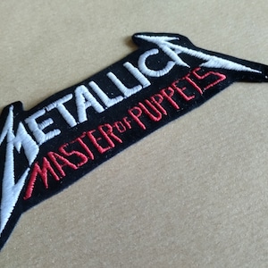 May include: Embroidered patch with the Metallica logo and the words "MASTER OF PUPPETS" in white and red on a black base. The logo is a stylized, angular design with a lightning bolt shape.
