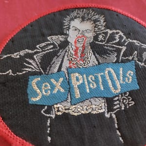 May include: Round black patch with a red border featuring a graphic of a man with blood dripping from his mouth. The words "Sex Pistols" are displayed on a blue banner with gold lettering. The patch is likely for clothing or accessories.