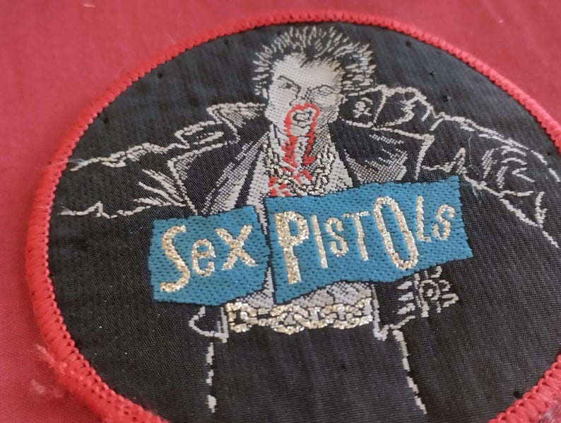 May include: Round black patch with a red border featuring a graphic of a man with blood dripping from his mouth. The words "Sex Pistols" are displayed on a blue banner with gold lettering. The patch is likely for clothing or accessories.