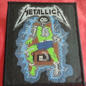 Könnte beinhalten: Schwarzer quadratischer Aufnäher mit dem Bandnamen "METALLICA" in Weiß, über einer Illustration eines grünen und blauen Skeletts auf einem elektrischen Stuhl. Blaue Blitze umgeben das Skelett. Der Aufnäher hat einen schwarzen Rand.