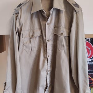 May include: A beige long-sleeved button-up shirt with two flap pockets and black shoulder patches.