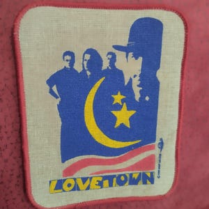 Può includere: Una toppa rettangolare con bordo rosso presenta una silhouette blu di una persona con un cappello, una mezzaluna e stelle. Sagome di tre persone sono a sinistra. La parola "LOVETOWN" è stampata in verde.