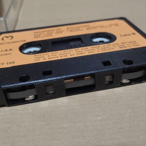 以下が含まれることがあります： オレンジ色のラベルが付いた黒いカセットテープ。ラベルには「Metalmaster」、「SJAE Stereo」、「MET 109」と書いてあります。テープには「Lato B」と表示されており、イタリア語で書かれたテキストがあります。