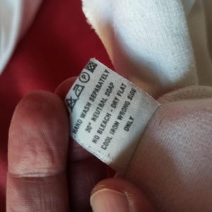 May include: A close-up of a white clothing label with washing instructions. The label states "HAND WASH SEPARATELY", "30&deg; NEUTRAL SOAP", "NO BLEACH - DRY FLAT", "COOL IRON WRONG SIDE", and "ONLY". The label is held by a person.