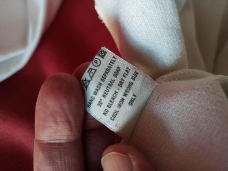 May include: A close-up of a white clothing label with washing instructions. The label states "HAND WASH SEPARATELY", "30&deg; NEUTRAL SOAP", "NO BLEACH - DRY FLAT", "COOL IRON WRONG SIDE", and "ONLY". The label is held by a person.