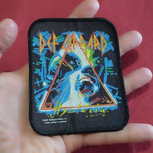 May include: A rectangular black fabric patch featuring the Def Leppard Hysteria album cover art. The design includes a central triangle, a stylised face, and blue and yellow geometric accents. The text reads Def Leppard and Hysteria.