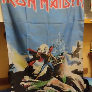 May include: A blue Iron Maiden flag with the band's red logo at the top. The flag showcases artwork from the "Phantom of the Opera" album, depicting a figure in a red cape against a fantasy backdrop.