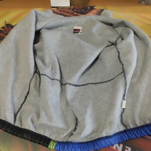 May include: A blue, green, and black jacket with a gray fleece lining. The jacket has a zipper closure and a drawstring hood.