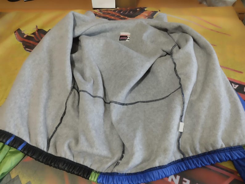 May include: A blue, green, and black jacket with a gray fleece lining. The jacket has a zipper closure and a drawstring hood.