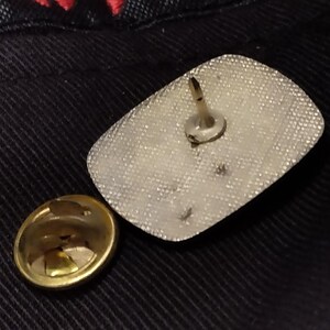 May include: The back of a silver pin with a white backing and a metal pin on the back.