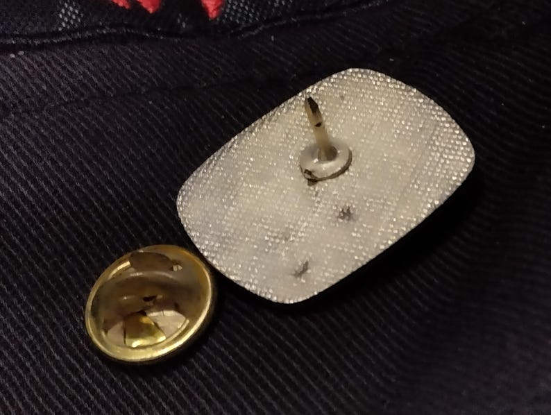 May include: The back of a silver pin with a white backing and a metal pin on the back.