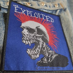 Pode incluir: Um patch azul com uma caveira com moicano vermelho e o texto "THE EXPLOITED" em branco. O patch é costurado em uma jaqueta jeans.