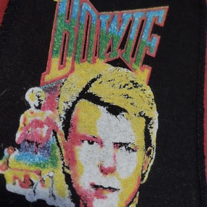 Può includere: Toppa nera con un'immagine colorata di David Bowie e il nome "BOWIE" in grandi lettere stilizzate. L'immagine ha un aspetto vintage, leggermente sbiadito, con un bordo rosso.