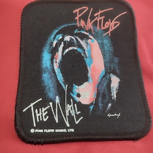 Könnte beinhalten: Ein rechteckiger schwarzer Stoffaufnäher mit einer Grafik eines schreienden Gesichts in Blau- und Rosatönen. Der Text Pink Floyd erscheint oben in Rosa, The Wall steht unten in Weiß, mit dem Text Pink Floyd Music Ltd. darunter.