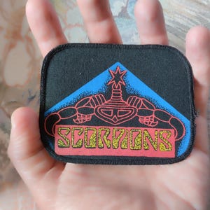 May include: A rectangular black fabric patch with rounded corners featuring a blue triangular graphic containing a red scorpion illustration and the word SCORPIONS in yellow block lettering on a red rectangular background at the bottom.