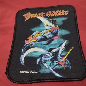 May include: A rectangular black fabric patch featuring the text Great White in yellow and red. The graphic depicts two sharks wearing sunglasses and playing electric guitars in shades of blue, yellow, and red against a dark background.