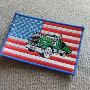 Può includere: Un camion semi-rimorchio verde con una cabina bianca e un rimorchio bianco è sovrapposto a una toppa di bandiera americana. La toppa ha un bordo blu.