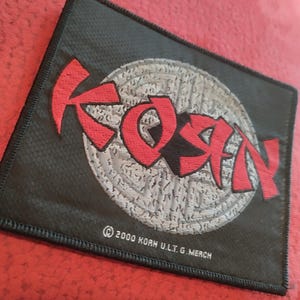 May include: Black rectangular patch with the band name "KORN" in red, stylized lettering. The letters are outlined in black. The background is a textured gray circle with a pattern. The patch has a black border and the text "© 2000 KORN U.L.T.G. MERCH".