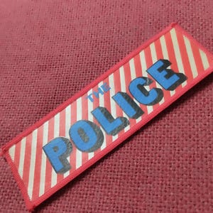 Może przedstawiać: Prostokątna naszywka z tkaniny z tłem w czerwono-białe ukośne paski. Napis THE POLICE jest wydrukowany pogrubionymi niebieskimi wielkimi literami z efektem czarnego cienia, wyśrodkowany na wzorze w paski znajdującym się na naszywce.