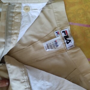 Puede incluir: Primer plano de pantalones Fila color beige, parcialmente desabrochados, mostrando el forro interior y la etiqueta de la marca. Los pantalones tienen un bot&oacute;n, cremallera y el logotipo de Fila. La tela parece ligera, adecuada para uso casual.