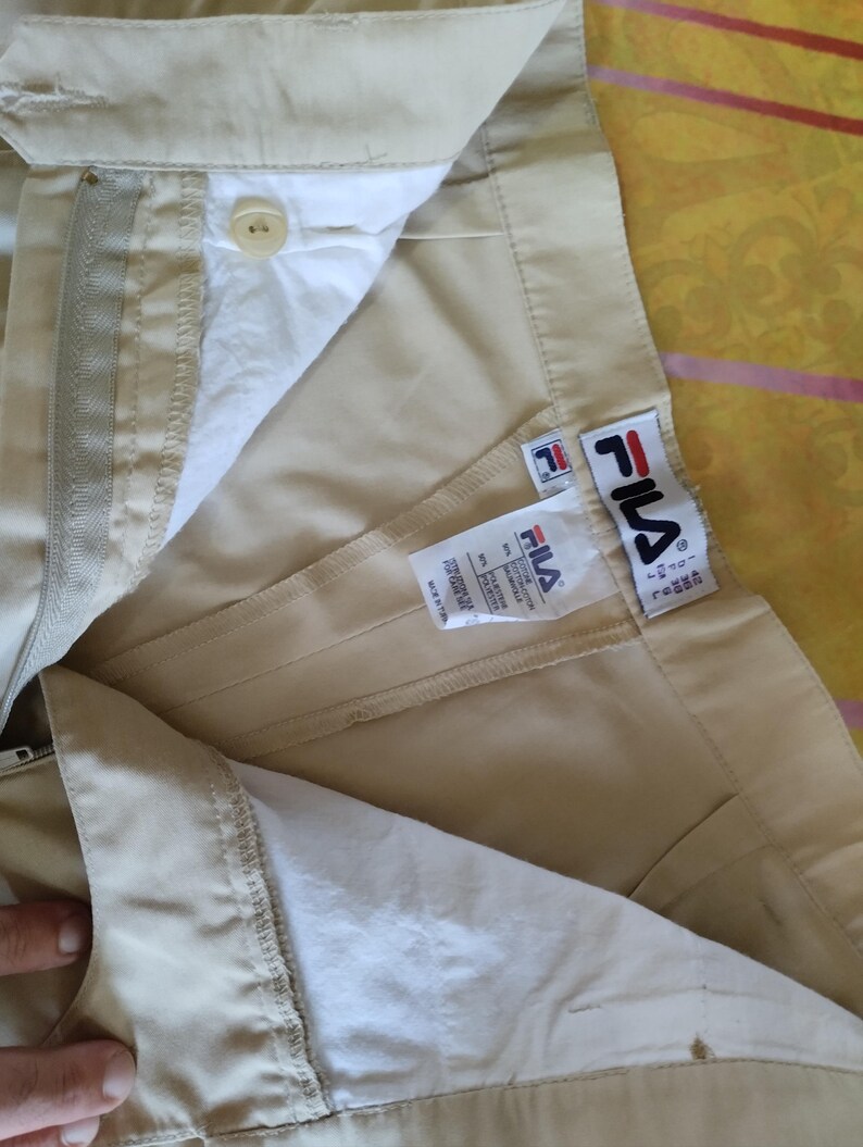 Puede incluir: Primer plano de pantalones Fila color beige, parcialmente desabrochados, mostrando el forro interior y la etiqueta de la marca. Los pantalones tienen un bot&oacute;n, cremallera y el logotipo de Fila. La tela parece ligera, adecuada para uso casual.
