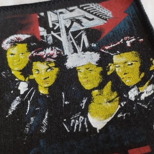 May include: A rectangular fabric patch with a black border featuring a graphic print of four individuals with yellow-toned skin and dark hair. Above them is a white architectural structure against a red background. The bottom edge shows partial red text.