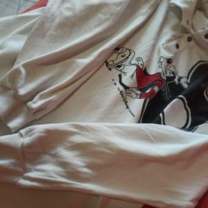 May include: Cream-coloured long-sleeved sweatshirt with a graphic of Disney's Goofy in a red and black outfit. The sweatshirt has a button-up collar and ribbed cuffs. The Disney Company copyright is visible.