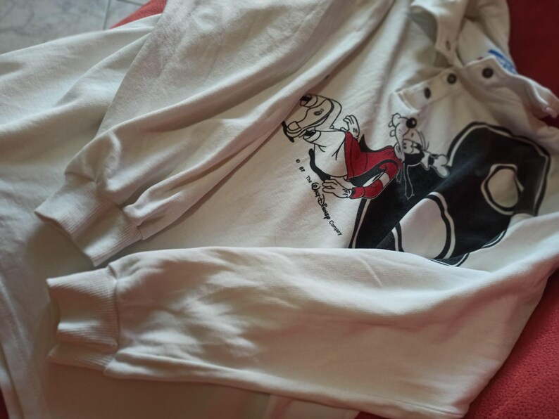 May include: Cream-coloured long-sleeved sweatshirt with a graphic of Disney's Goofy in a red and black outfit. The sweatshirt has a button-up collar and ribbed cuffs. The Disney Company copyright is visible.