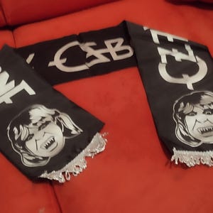 Puede incluir: Bufanda negra con texto blanco y una imagen de dibujos animados de una cara con colmillos. La bufanda tiene el texto "NEE" y "CEE" en los extremos, y "CSB" en el medio. Flecos plateados en los extremos.