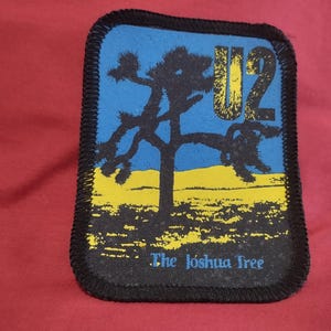 Könnte beinhalten: Ein rechteckiger Stoffaufnäher mit schwarzem, gesticktem Rand. Das Design zeigt einen blauen Himmel, eine gelbe Wüstenlandschaft und die schwarze Silhouette eines Joshua-Baums. Der Text U2 steht gelb oben rechts, The Joshua Tree ist unten blau aufgedruckt.