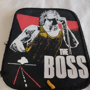 Puede incluir: Un parche de tela rectangular con borde negro que presenta el gráfico de un cantante con camiseta amarilla sosteniendo un micrófono. El diseño incluye un fondo triangular rojo, un gráfico de carretera y el texto blanco THE BOSS impreso en la parte inferior.