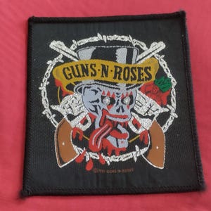 May include: Black square patch with the Guns N' Roses logo. The design features a skull in a top hat, crossed revolvers, and a banner with the band's name. The patch is edged with barbed wire.