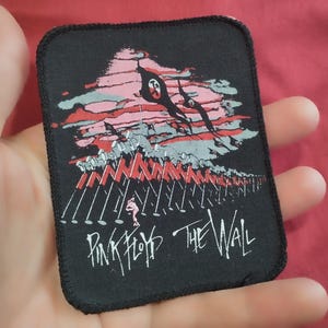 Peut inclure: Un écusson rectangulaire en tissu noir présentant une illustration graphique de marteaux en marche et un ciel rose. Le motif inclut le texte Pink Floyd The Wall en lettres blanches en bas. L'écusson a une bordure cousue noire.