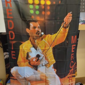 May include: A black and red fabric flag featuring a photo of Freddie Mercury performing on stage. The flag has the words "FREDDIE" and "MERCURY" printed vertically on the left and right sides of the image.