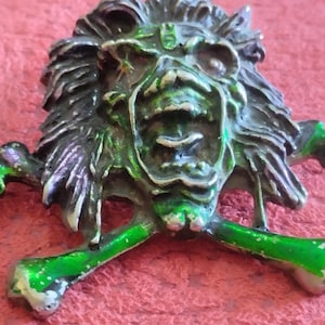Könnte beinhalten: Ein metallener Totenkopf mit grünen und silbernen Akzenten, mit gekreuzten Knochen. Der Totenkopf hat ein detailliertes, strukturiertes Design vor einem roten Hintergrund. Das Metall hat ein gealtertes Aussehen.