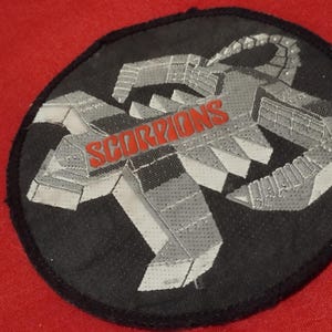 Könnte beinhalten: Ein schwarzer, kreisförmiger Aufnäher mit einem grauen Skorpion-Design und dem Wort "SCORPIONS" in Rot. Der Aufnäher ist wahrscheinlich ein Emblem oder Logo, möglicherweise für eine Band oder ein Team.
