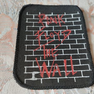 Könnte beinhalten: Ein rechteckiger Stoffaufnäher mit schwarzem Hintergrund, einem weißen Ziegelsteinmuster und dem roten Schriftzug Pink Floyd The Wall. Der Aufnäher hat einen schwarz umkettelten Rand und abgerundete Ecken im ikonischen Albumdesign.