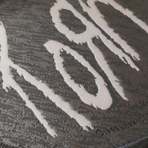 Pode incluir: Close-up de um patch preto e cinza texturizado com a palavra "KORN" em branco, com letras estilizadas em pinceladas. O patch tem uma borda azul escura.