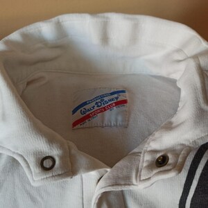 May include: A close-up of a white collared shirt with a button-down closure. The shirt features a tag with the text "Walt Disney Sports Club" and red and blue accents. The shirt has a black and white stripe design on the side.