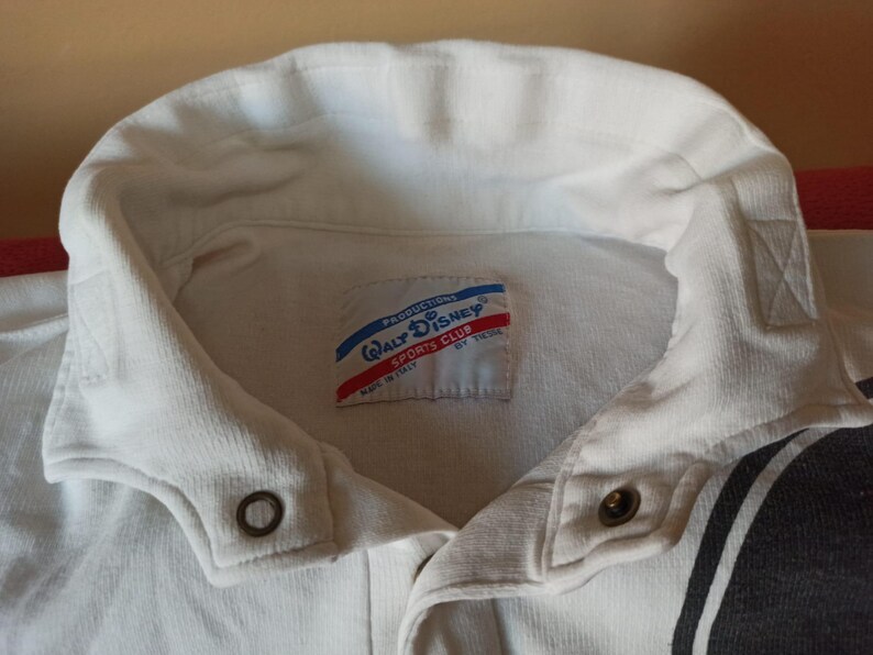 May include: A close-up of a white collared shirt with a button-down closure. The shirt features a tag with the text "Walt Disney Sports Club" and red and blue accents. The shirt has a black and white stripe design on the side.